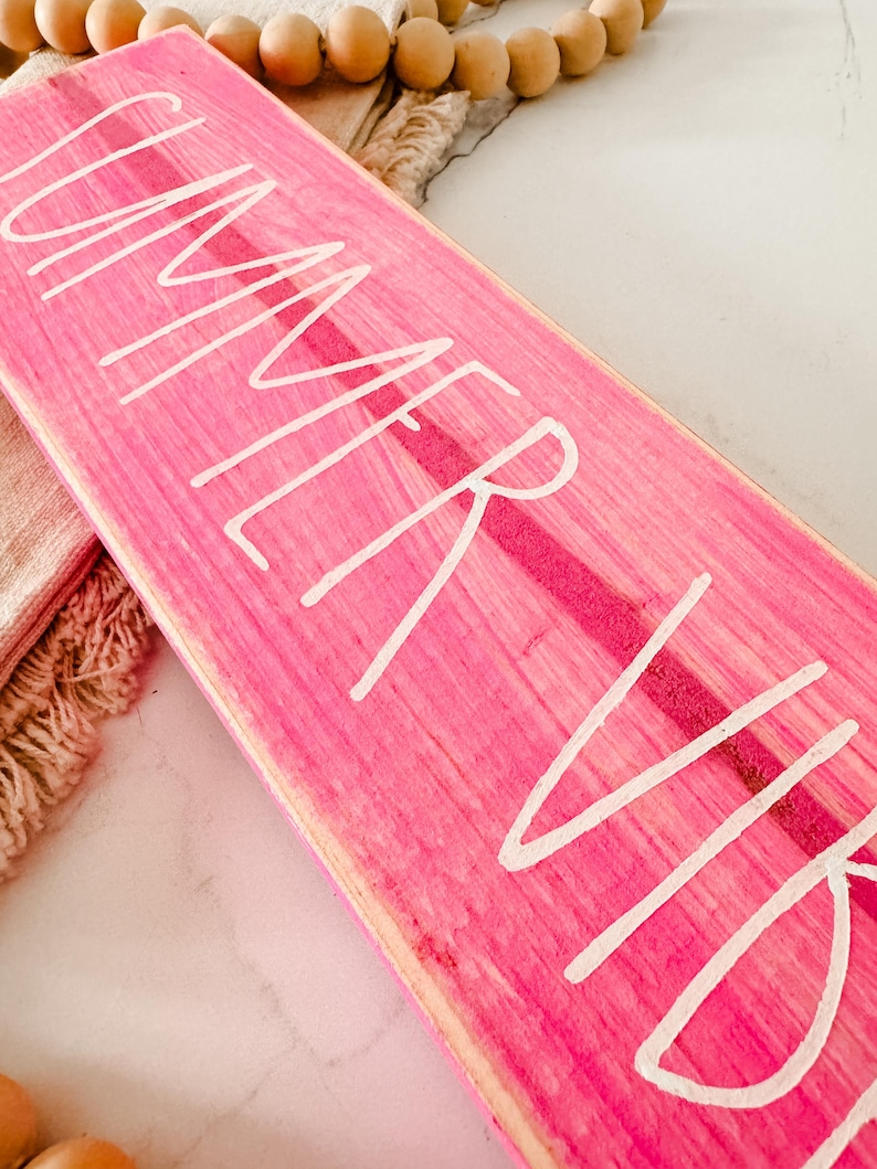 May include: A rectangular wooden sign painted in a vibrant hot pink colour with a visible wood grain texture. The text "SUMMER VIBES" is written across the surface in a white, hand-lettered script font. The sign has a flat, elongated shape.