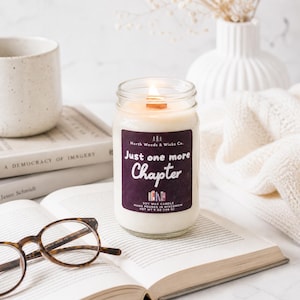 K&ouml;nnte beinhalten: Eine brennende Sojawachskerze in einem Glas mit Holzdocht, beschriftet mit "Just one more Chapter". Die Kerze steht auf einem offenen Buch mit Brille, in der N&auml;he einer Tasse und eines B&uuml;cherstapels. Nettogewicht 198 g.