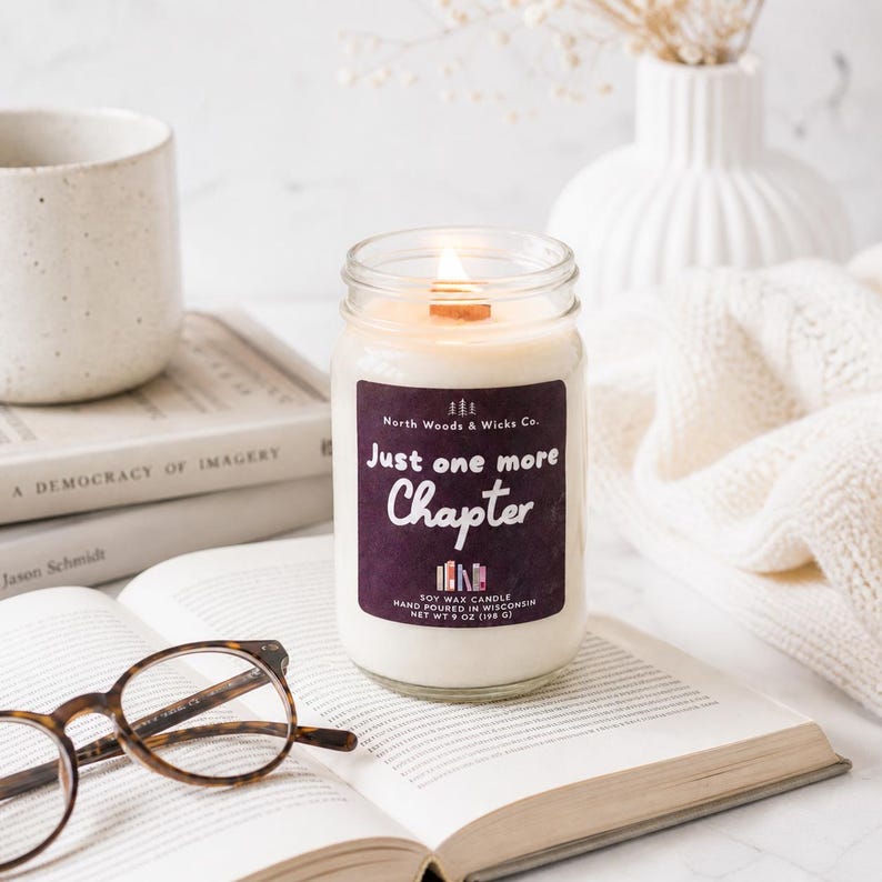 K&ouml;nnte beinhalten: Eine brennende Sojawachskerze in einem Glas mit Holzdocht, beschriftet mit "Just one more Chapter". Die Kerze steht auf einem offenen Buch mit Brille, in der N&auml;he einer Tasse und eines B&uuml;cherstapels. Nettogewicht 198 g.
