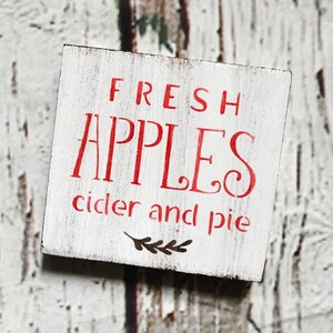 May include: A small, square, white wooden sign with red lettering. The sign reads "FRESH APPLES cider and pie" in a decorative font. A small sprig of leaves is below the text. The wood has a distressed, rustic appearance.