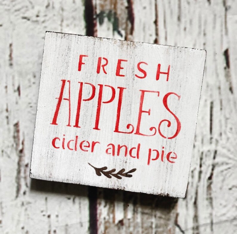 May include: A small, square, white wooden sign with red lettering. The sign reads "FRESH APPLES cider and pie" in a decorative font. A small sprig of leaves is below the text. The wood has a distressed, rustic appearance.