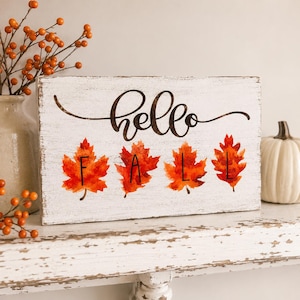 May include: A rectangular white wooden sign with a distressed finish featuring the black cursive text hello and four vibrant orange and red watercolor-style maple leaves, each marked with a single black letter to spell out the word FALL.