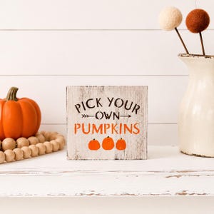 May include: A square wooden sign with a distressed white finish featuring the black text PICK YOUR OWN and orange text PUMPKINS, accented by an arrow graphic and three small orange pumpkin illustrations centered at the bottom of the sign.