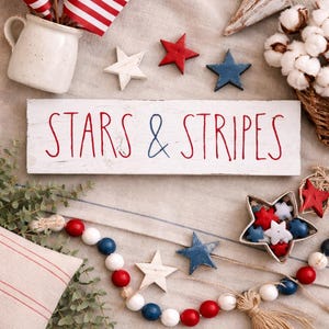 Puede incluir: Un letrero rectangular de madera blanca con el texto STARS & STRIPES en rojo y azul, acompa&ntilde;ado de peque&ntilde;as banderas estadounidenses en una jarra blanca, estrellas de madera rojas y azules, un cuenco en forma de estrella y una guirnalda de cuentas.