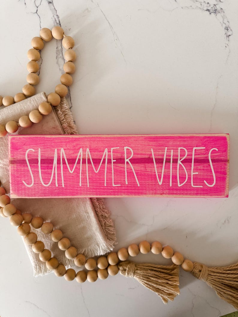 May include: A rectangular wooden sign painted in a bright hot pink with the text "SUMMER VIBES" written in white, sans-serif capital letters. The sign is accompanied by a strand of natural wooden beads and a beige fabric tassel.