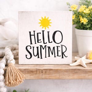 May include: A white wooden sign with the text "HELLO SUMMER" in black and a yellow sun illustration. The sign sits on a wooden shelf, alongside a starfish and a potted plant, evoking a summer theme.