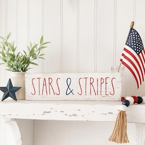 Puede incluir: Un letrero rectangular de madera blanca con bordes desgastados que presenta el texto en rojo y azul STARS & STRIPES. El letrero se muestra junto a una peque&ntilde;a bandera estadounidense, una estrella de metal azul y una cadena de cuentas de madera rojas, blancas y azules.