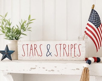 May include: A rectangular white wooden block sign with distressed edges featuring the red and blue text STARS & STRIPES. The sign is displayed alongside a small American flag, a blue metal star, and a string of red, white, and blue wooden beads.