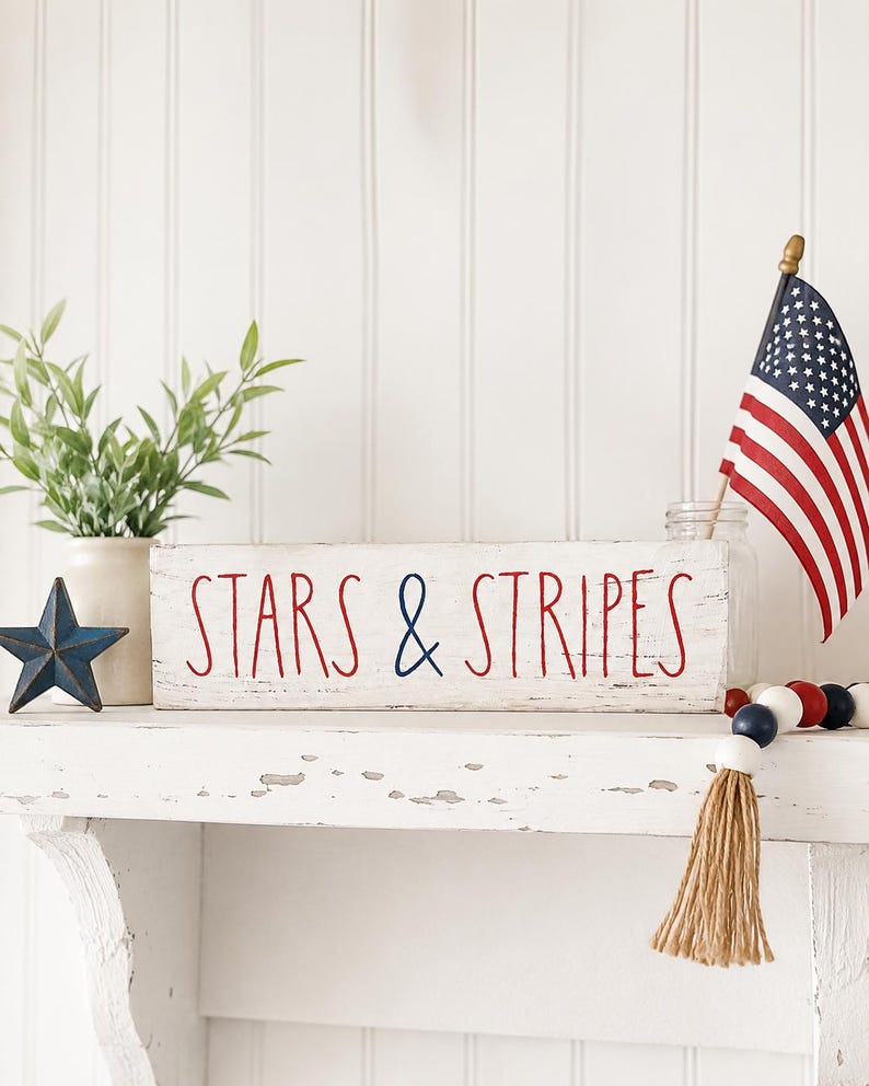 Puede incluir: Un letrero rectangular de madera blanca con bordes desgastados que presenta el texto en rojo y azul STARS & STRIPES. El letrero se muestra junto a una peque&ntilde;a bandera estadounidense, una estrella de metal azul y una cadena de cuentas de madera rojas, blancas y azules.