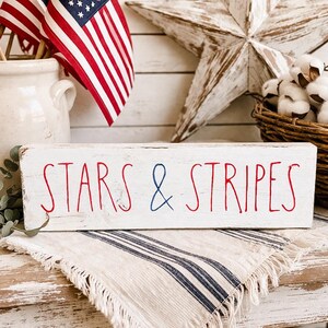 Puede incluir: Un letrero rectangular de madera blanca con bordes desgastados que presenta el texto STARS & STRIPES en letras rojas y azules. La palabra STARS y la palabra STRIPES est&aacute;n escritas en rojo, mientras que el s&iacute;mbolo ampersand est&aacute; en azul.