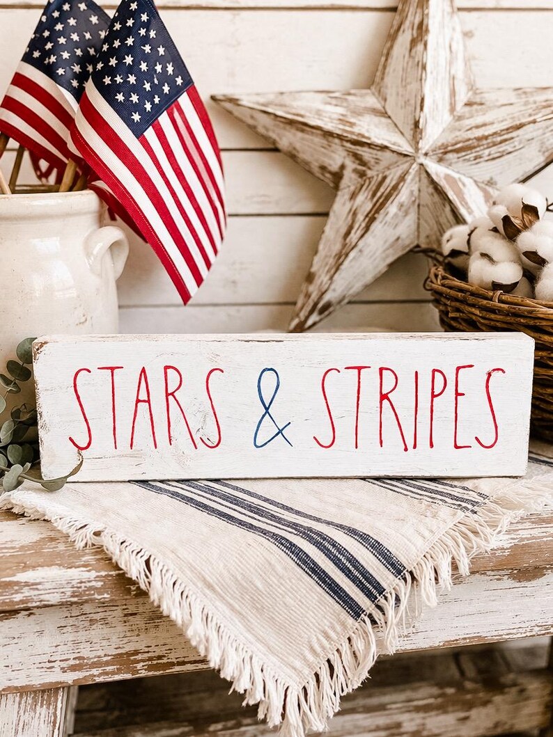 Puede incluir: Un letrero rectangular de madera blanca con bordes desgastados que presenta el texto STARS & STRIPES en letras rojas y azules. La palabra STARS y la palabra STRIPES est&aacute;n escritas en rojo, mientras que el s&iacute;mbolo ampersand est&aacute; en azul.