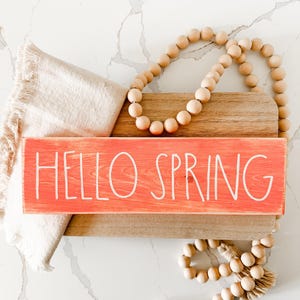 May include: A wooden sign with the words "HELLO SPRING" painted in white on a coral pink background. The sign is sitting on a wooden chopping board with a strand of wooden beads draped over it.