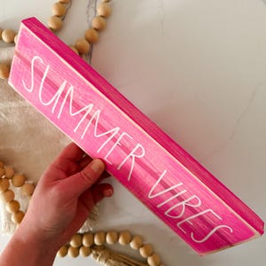 May include: A rectangular wooden sign painted in a bright hot pink colour with the text SUMMER VIBES written in white, sans-serif capital letters. The sign is accompanied by a decorative strand of natural light-coloured wooden beads with tassels.