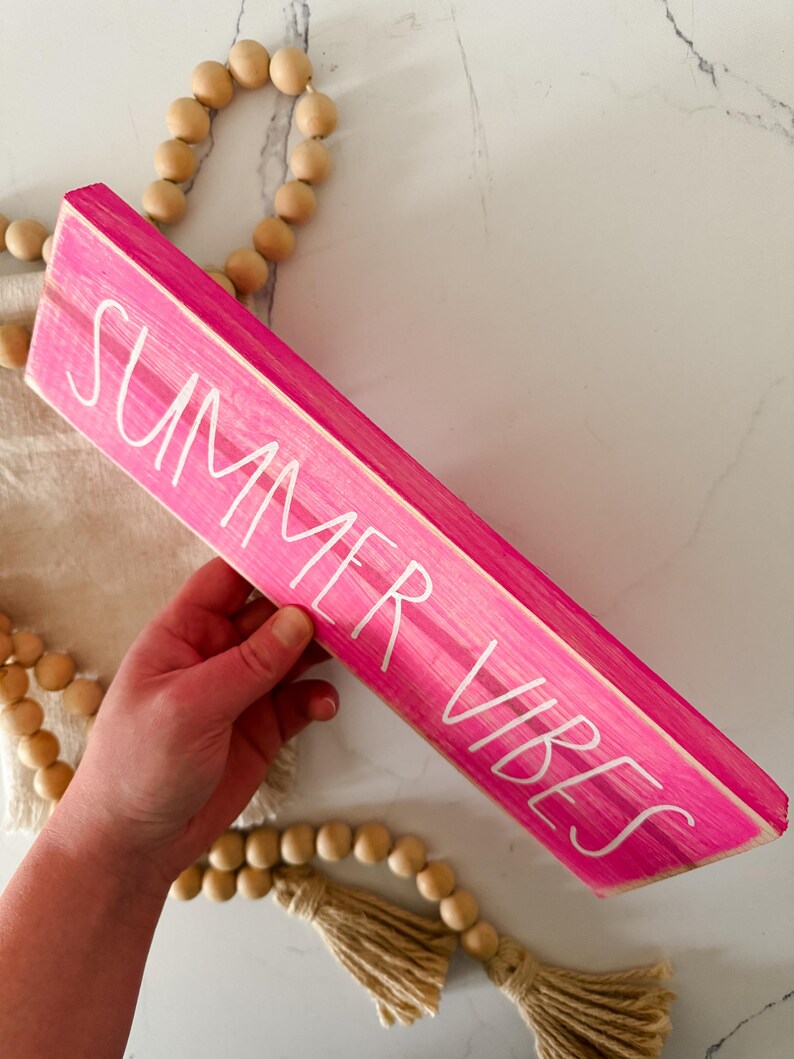 May include: A rectangular wooden sign painted in a bright hot pink colour with the text SUMMER VIBES written in white, sans-serif capital letters. The sign is accompanied by a decorative strand of natural light-coloured wooden beads with tassels.