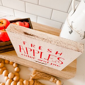 May include: A rustic wooden sign with the words "FRESH APPLES cider and pie" in red lettering. The sign is on a wooden shelf, with a crate of red apples and a white pitcher in the background. Wooden beads and a jute tassel are in the foreground.