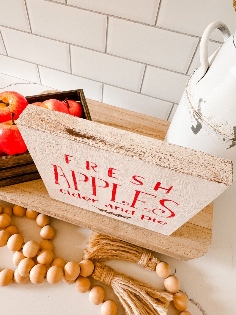May include: A rustic wooden sign with the words "FRESH APPLES cider and pie" in red lettering. The sign is on a wooden shelf, with a crate of red apples and a white pitcher in the background. Wooden beads and a jute tassel are in the foreground.