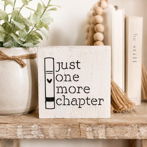 Puede incluir: Un bloque de madera blanco envejecido con la frase "just one more chapter" en letras negras. Una ilustración negra de un libro está a la izquierda. El bloque está sobre una estantería de madera, con libros y una planta en el fondo.