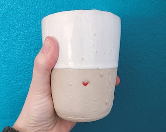May include: A white ceramic mug with a beige bottom and a small red heart design. The mug is being held by a hand.