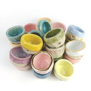Könnte beinhalten: Eine Sammlung kleiner, runder Keramikschalen in verschiedenen Pastell- und leuchtenden Farben wie Rosa, Gelb, Blau, Grün und Weiß. Jede Schale hat ein glatt glasiertes Inneres und einen strukturierten, unglasierten Boden, einige mit einem kleinen Herz.