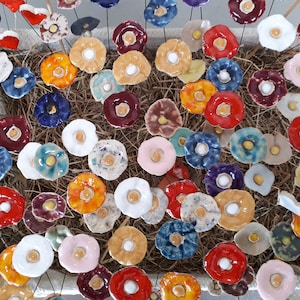 Op de afbeelding: Een verzameling keramische bloemenversieringen in verschillende kleuren, waaronder rood, blauw, geel en wit. De bloemen zijn gerangschikt in een mand gevuld met stro.