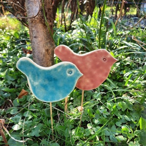 May include: Two ceramic bird figurines, one blue and one pink, are placed in a garden setting. The birds are perched on wooden sticks and have a whimsical, decorative style.