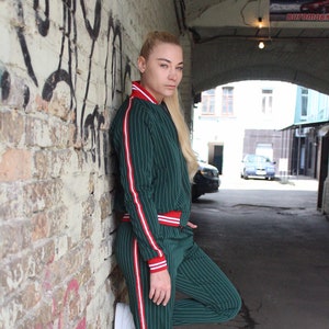 May include: A woman wearing a green and white striped tracksuit with red accents. She is standing against a brick wall and has her leg bent at the knee. She is wearing white sneakers.