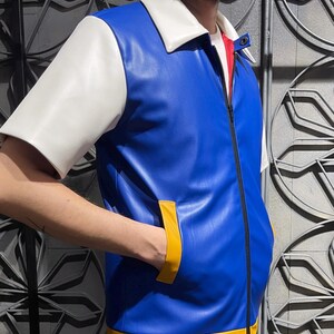May include: A short-sleeved jacket made of blue faux leather with a white collar and sleeves. The jacket features a black front zipper, yellow trim along the bottom hem, and yellow accents on the two side pockets. A red inner lining is visible.