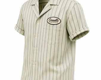 Puede incluir: Una camisa de manga corta con botones en color crema claro con rayas verticales de color verde salvia. La camisa presenta un cuello tipo camp, un bolsillo en el pecho y un parche ovalado marrón en el bolsillo con el texto Cassell's CONFIDENCE, STYLE & SUCCESS.