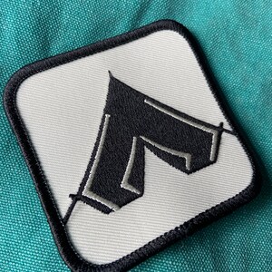 May include: A white square patch with black stitching around the edges. A black tent with white stitching is embroidered on the patch.