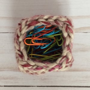 May include: A small, crocheted basket with a red and white pattern is filled with colorful paper clips.