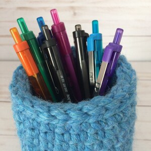 May include: A blue crocheted pencil holder filled with colorful pens. The pens are a variety of colors, including orange, green, blue, purple, and black.