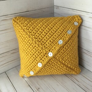 May include: A square mustard yellow crochet pillow featuring a diagonal seam across the front. The seam is embellished with a row of seven round white buttons, each with four holes, arranged in a straight line from the top right to the bottom left.