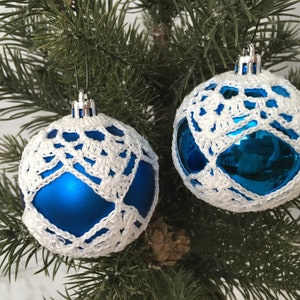 May include: Two spherical Christmas ornaments featuring a bright blue base covered in a delicate white crocheted lace overlay. Each ornament has a silver-toned cap at the top with a small loop for hanging, suspended from the branches of a pine tree.