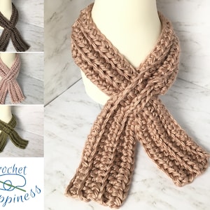 May include: A crocheted scarf in a light brown color. The scarf is made with a textured stitch and is shown folded and tied in a knot. The scarf is displayed on a white mannequin.