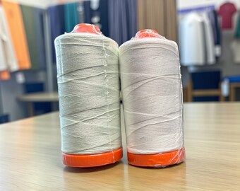 May include: Two spools of white thread with orange bases sit on a wooden table. The thread is tightly wound around the spools, and the orange bases are visible at the bottom. The background shows clothing and tables.