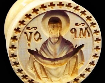 May include: A light-colored, wooden plug with a carved religious icon. The icon depicts a figure with a halo, arms outstretched, and text in a circular frame with cross-shaped designs. The plug is cylindrical.