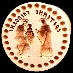 May include: A round, light brown wooden carving with religious figures and text. The carving features two figures and a child, surrounded by a circle of small holes. The text is in a foreign language.