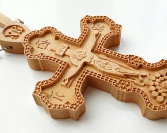 May include: A detailed wooden Orthodox cross with intricate carvings, including a depiction of Jesus Christ. The cross is light brown and features decorative patterns and religious symbols. It is connected to a wooden chain.