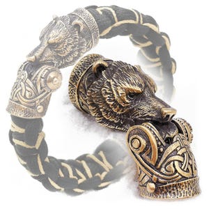 Puede incluir: Un brazalete de cuero trenzado negro y crema con un dije de cabeza de oso de bronce. La cabeza del oso está detallada y tiene una expresión feroz. El brazalete es una excelente manera de mostrar tu amor por la naturaleza y el aire libre.