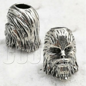 Puede incluir: Dos llaveros de metal plateado, uno con forma de cabeza humana con una cara peluda y el otro con forma de cabello.