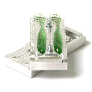 May include: A two-part aluminum fishing lure mold containing two translucent green soft plastic baits with red glitter flecks. The mold features precision-cut cavities for shaping the lures, with two metal alignment pins visible on the top surface.