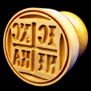 May include: A light-colored wooden stamp with carved Cyrillic letters within a circular frame. The stamp has a rounded handle and is set against a black background. The letters are deeply carved into the wood.