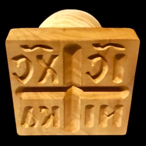 May include: A light brown wooden stamp with carved symbols and a rounded wooden handle. The stamp has a rectangular shape with a series of carved symbols. The handle is a light brown, rounded shape. The background is black.