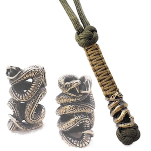 以下が含まれることがあります： 緑と茶色のパラコードに、それぞれ異なるデザインの真鍮製の蛇のビーズが3つあります。ビーズは鱗と牙で装飾されています。