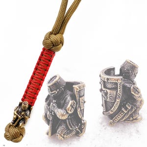 May include: A bronze-coloured metal bead featuring a detailed depiction of a kneeling knight in armour holding a sword and shield. The bead is designed to be used as a fidget toy or a decorative element on a keychain or lanyard.