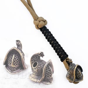 May include: Two bronze gladiator helmet beads with intricate details, attached to a brown and black paracord lanyard.