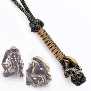 Peut inclure: Ensemble de perles et de lanières sur le thème des extraterrestres en bronze. La lanière est composée d'un cordon tressé beige et noir, d'un nœud décoratif et d'une perle en forme de tête d'Alien en bronze. Deux perles Alien supplémentaires sont également présentées.