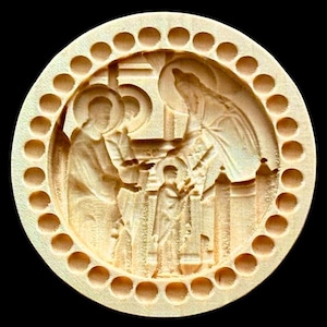Puede incluir: Una talla de madera circular y de color claro que representa una escena religiosa. La talla presenta figuras con halos, una cruz y elementos arquitectónicos. El borde exterior tiene una serie de hendiduras circulares espaciadas uniformemente.