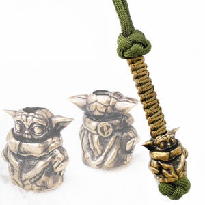 Puede incluir: Un brazalete de paracord verde y marrón con un dije de latón que representa a un personaje de ficción popular. El dije está unido al brazalete con un nudo.