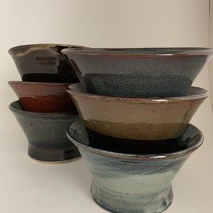 Puede incluir: Un juego de seis cuencos de cerámica en varios tonos de marrón, negro y azul. Los cuencos están apilados uno encima del otro, con el cuenco más grande en la parte inferior y el más pequeño en la parte superior.